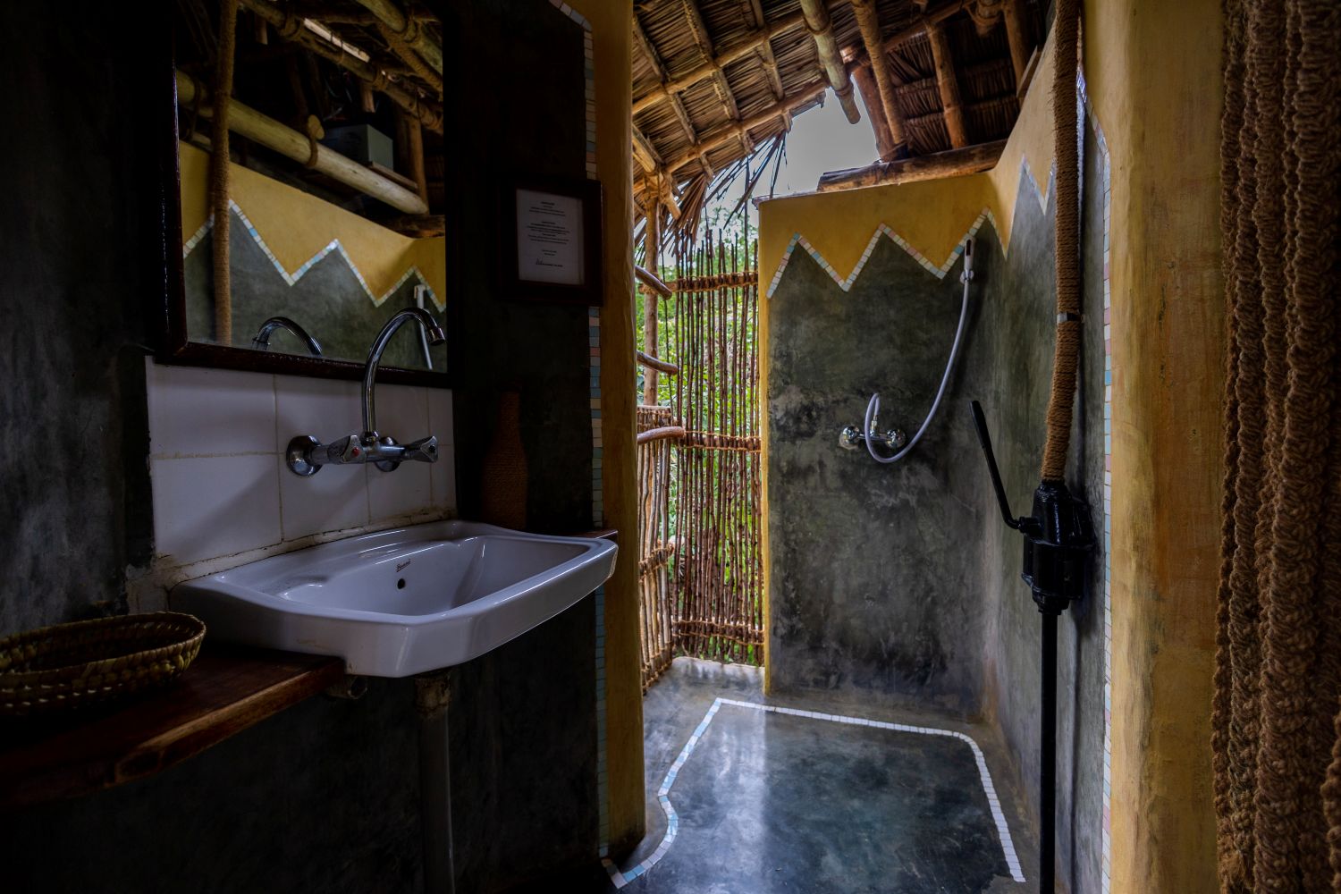 A bungalow bathroom finished in polished grey and ochre render, with a wall-mounted basin, a bare compost-toilet cubicle and an open shower whose only screen is a panel of vertical reeds onto the forest.