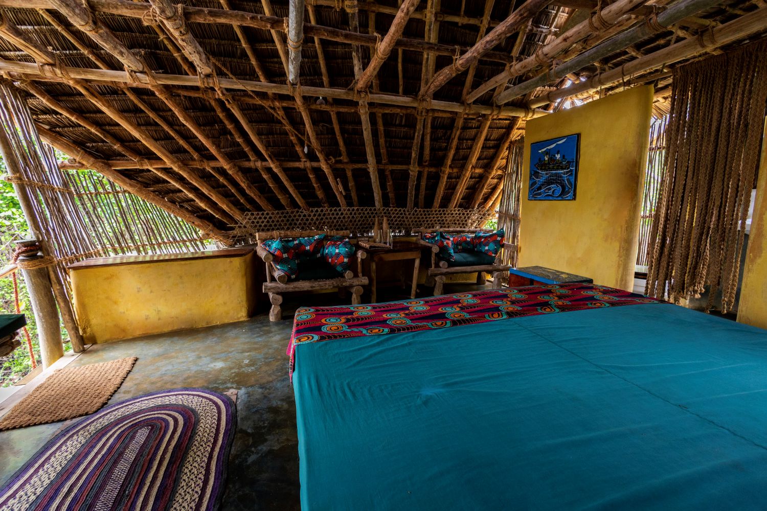 Inside a bungalow, a low bed dressed in teal cotton with a patterned throw, facing carved timber armchairs under an open ceiling of mangrove poles and thatch, with woven reed screens instead of window glass.